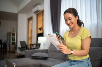 Asian woman sitting on sofa with mobile phone paying bills at home