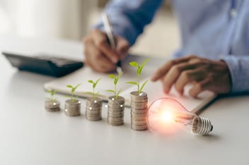 energy saving ideas and money saving or investment Businessman holding a light bulb and a pile of coins on a table with a notebook while taking notes on a notebook with piles of coins on the table