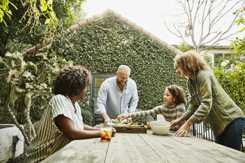 Father bringing try of food to family sitting at picnic table in backyard