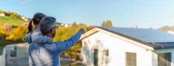 Rear view of dad holding her little girl in arms and showing at their house with installed solar panels. Alternative energy, saving resources and sustainable lifestyle concept.