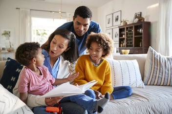 Young mother sitting on sofa in the living room with her daughter beside her and toddler son on her knee, father standing behind them, close up