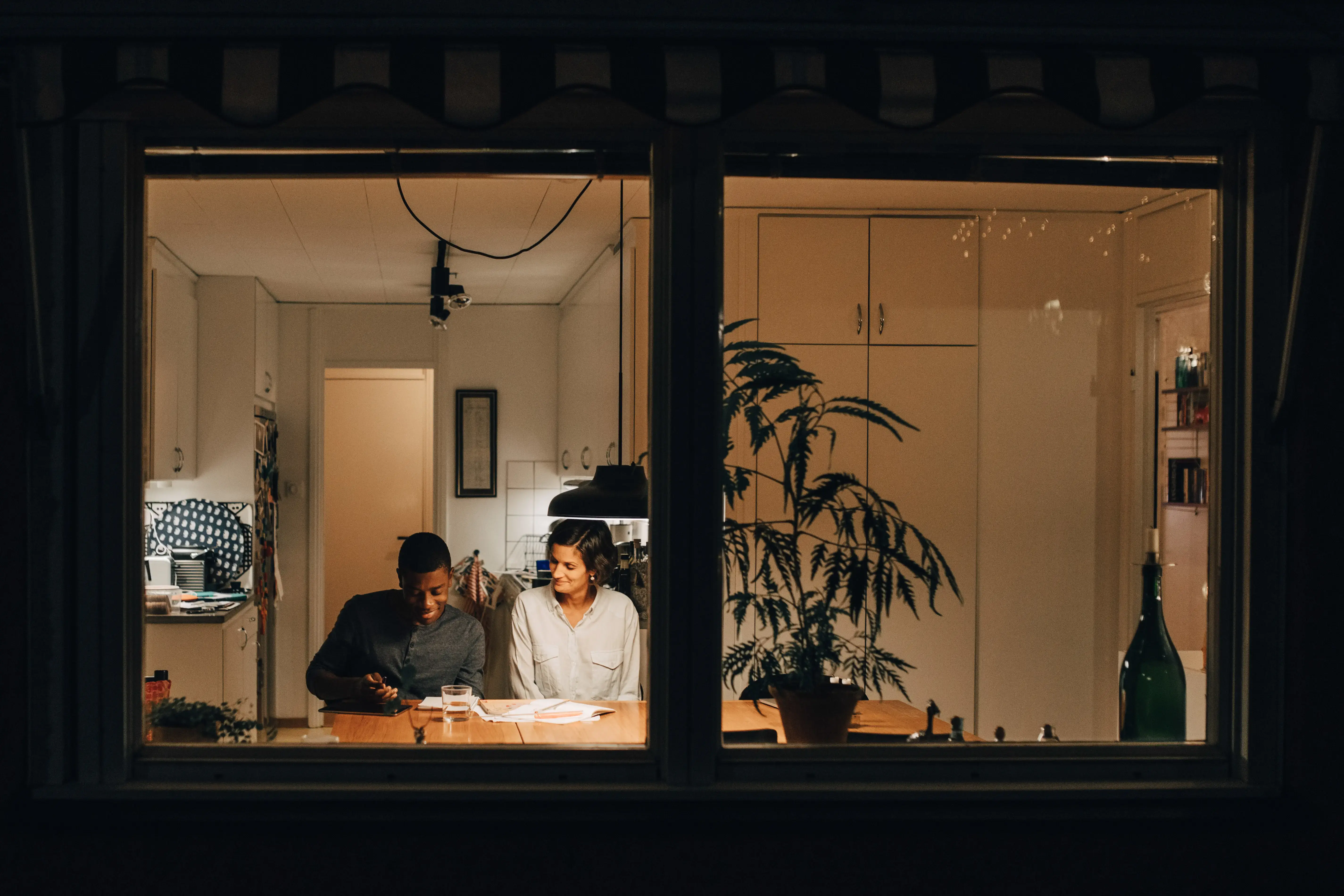 Mother assisting son in studying at home seen through window