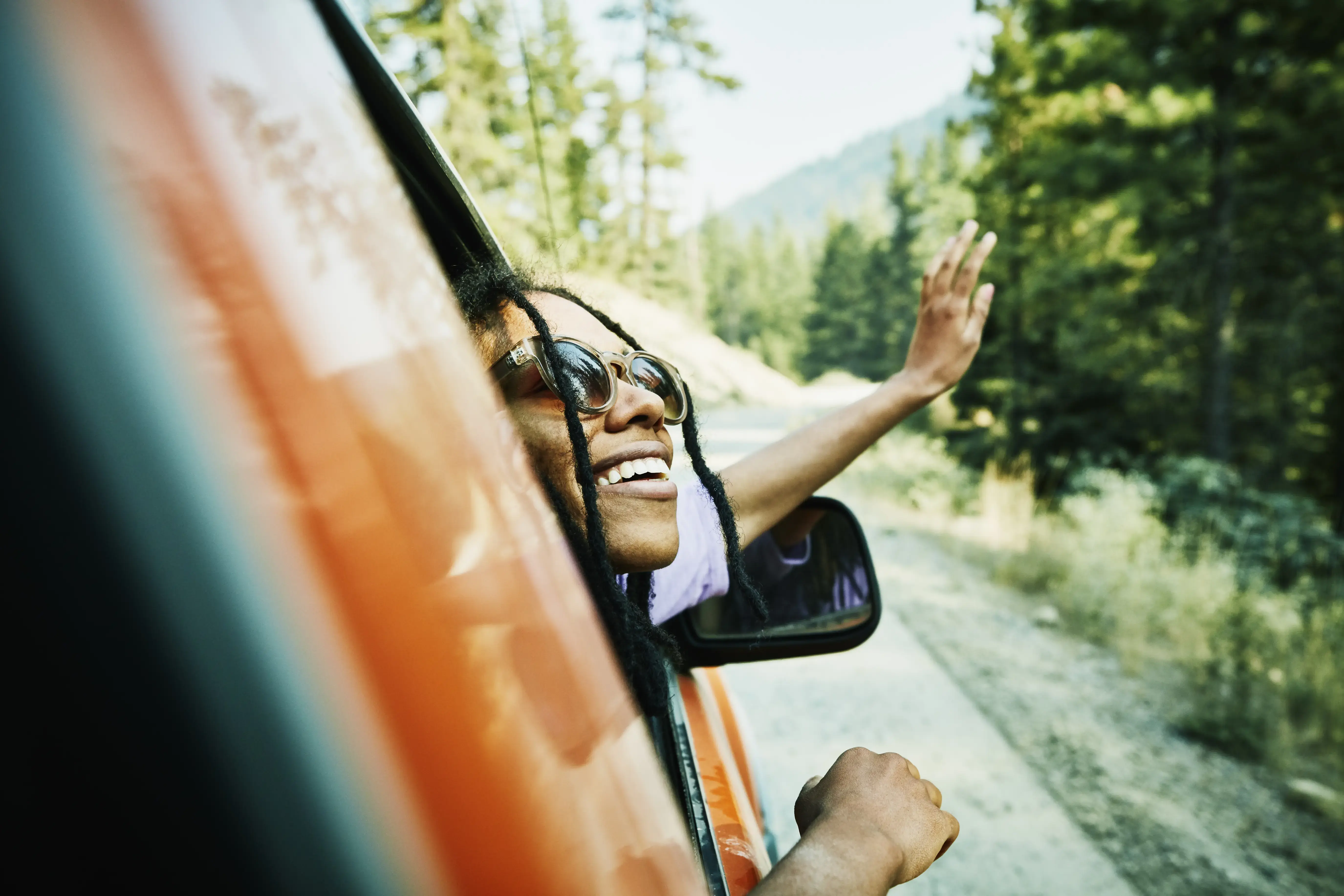 Smiling woman with head and hand out of car window enjoying view of mountains