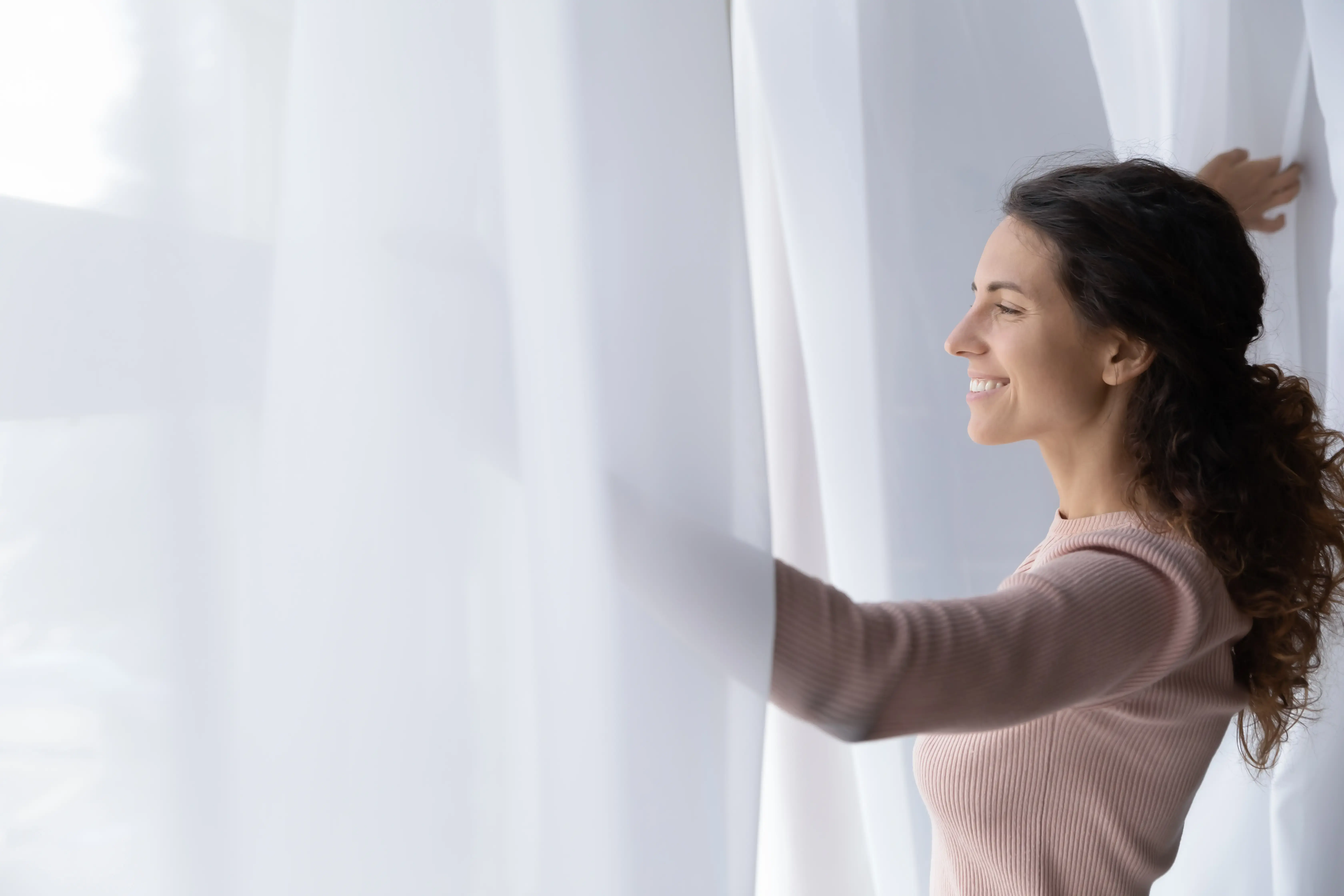 Young Latina woman open curtains looks out window feels happy